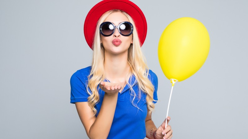 Pretty-young-woman-in-sunglasses-with-air-balloon-sends-an-air-kiss-over-gray-background-845194082_2